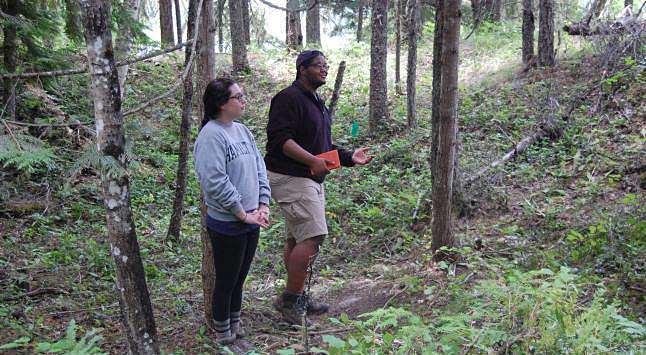 LEMON CREEK — Archaeology students Bridget Maguire and Kevin Castro stand in the indentation left by an old pithouse. They explained that 30 to 50 cubic meters of soil would have had to have been removed to build a pit house. The digging would have been done by women using digging sticks and the job would have been completed in about a week.  Laura Stovel photo