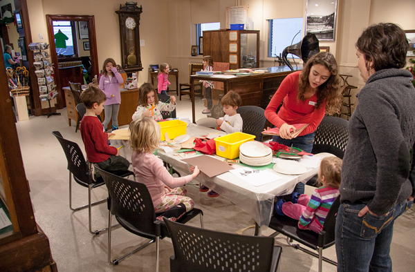 A different group of children checks out the Christmas Plates decorating table, helped by Hailey, another Museum host (in red) as one of the moms looks on. Jason Portras photo